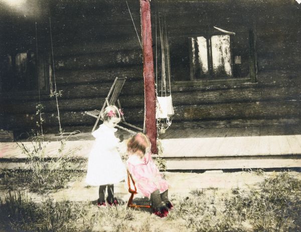 Jeannette Holt is standing and getting the tangles out of her brother Alfred Holt's hair in front of Island Lodge porch. Alfred is sitting in a small chair. Caption reads: "Jeannette and Alfred — Getting the Tangles Out."