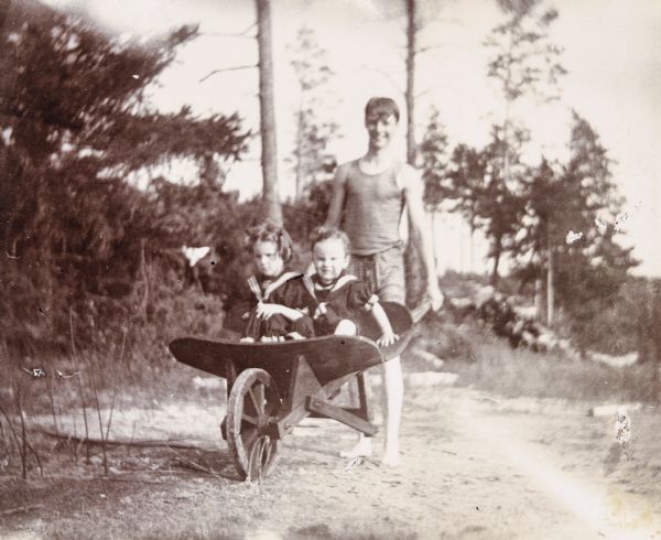 Smiling Uncle Wallace Rumsey is pushing a wooden wheelbarrow with Jeannette and Alfred Holt inside. They are on the dirt path leading to the swimming beach on the shore of Archibald Lake. They are all wearing bathing suits. Caption reads: "Fun on the beach — Alfred and Jeannette — Uncle Wallace."