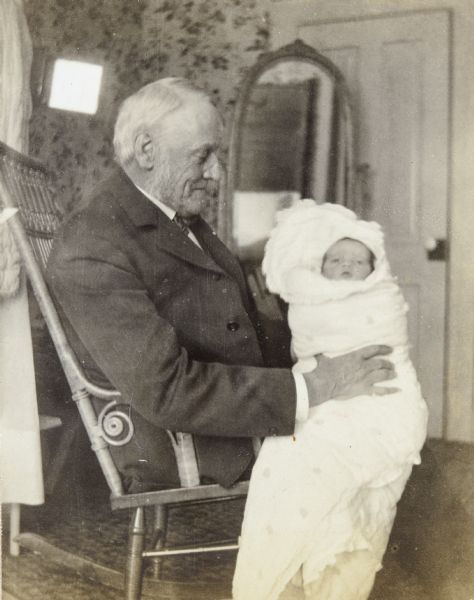Sitting in a wicker rocking chair at the Holt family home, Grandfather I.P. Rumsey is holding baby granddaughter Eleanor Holt who is swaddled in a blanket. A full-length mirror is standing in the background. Caption reads: "Grandfather and Eleanor."