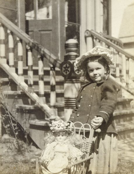 Jeannette Holt is posing with her doll who is sitting in a wicker baby carriage. In the background are the stairs leading up to the Holt family home. Caption reads: "Jeannette's 5th Birthday - May 8, 1901."