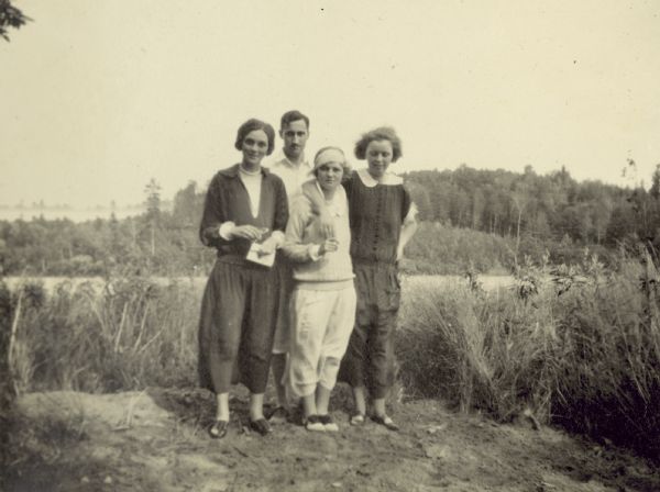 Standing together on a windy day (L to R) are: Blanche Unknown, Gordon Wheeler, Lillian Wheeler and Jane MacIdorney(?), with her arm around Lillian Wheeler's shoulder. Archibald Lake is in the background.

Caption reads: "Blanche, Gordon, Lillian, Jane MacIdorney (?)." Page title reads: "Archibald Lake, August 1923."