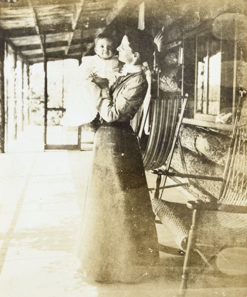 A woman wearing a long skirt and blouse is standing and holding baby Eleanor Holt in her arms while standing on the front porch of Island Lodge on Archibald Lake. Eleanor is looking at the camera.