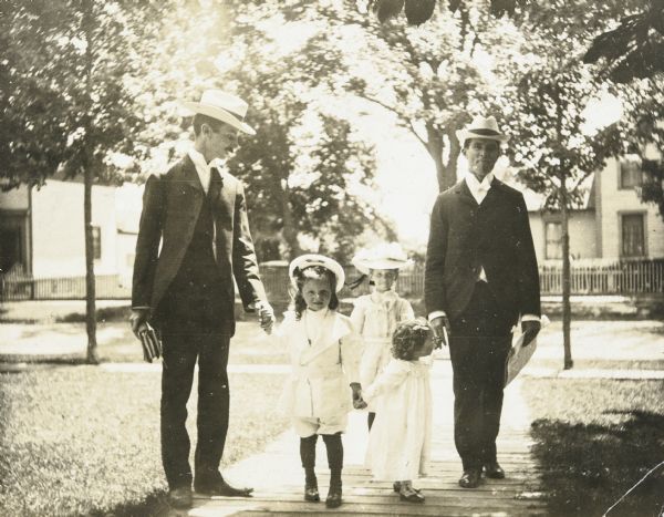 W.A. Holt and Rev. John Robertson Macartney are escorting the Holt children home from Sunday School. Names from left to right: W.A. Holt, Alfred Holt, Jeannette Holt, Eleanor Holt, and Rev. Macartney. W.A. Holt is holding a couple of books. Rev. Macartney is holding a newspaper. The group is standing on the wooden walkway that leads to the Holt family home. Oconto's Main Street is in the background. Caption reads: "Returning from Sunday School" and identifies Father and Rev. J.R. Macartney.
