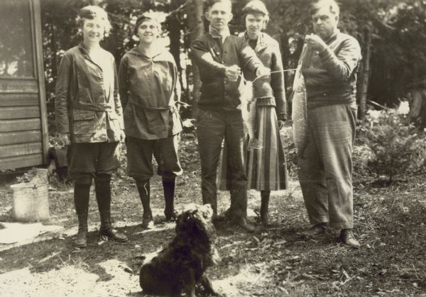 In the foreground, the Holt family dog, an English Cocker Spaniel, is looking up at freshly-caught fish on a stringer held by a man standing behind with a group of three women and one man. Lucy Rumsey Holt is the first person on the left. A portion of a screened porch is visible. Tall trees and Archibald Lake are in the background.