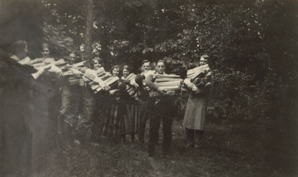 A group of people lined up in a row are carrying arm loads of split pine logs. Left to right: Unknown, Jeannette Holt, Unknown, Unknown, Eleanor Holt, Unknown, Unknown, Donald Holt, and W.A. Holt. Caption reads: "A Rainy Day Occupation." 