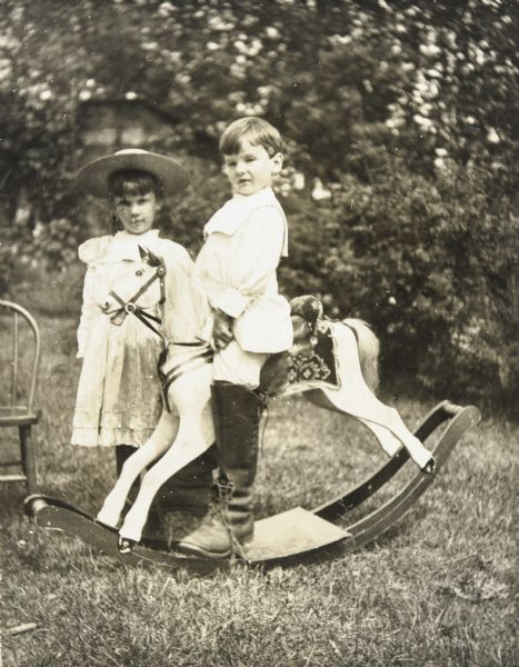 A portrait in the backyard of the Holt family home of Alfred Holt on the day he got his curls cut. Sitting on the rocking horse, Alfred is wearing a leather boot which is much too big for him. Looking at the camera, Jeannette Holt is standing tall, next to a wooden chair on the lawn on the left. Caption reads: "Don't I look like father," and identifies Jeannette and Alfred.