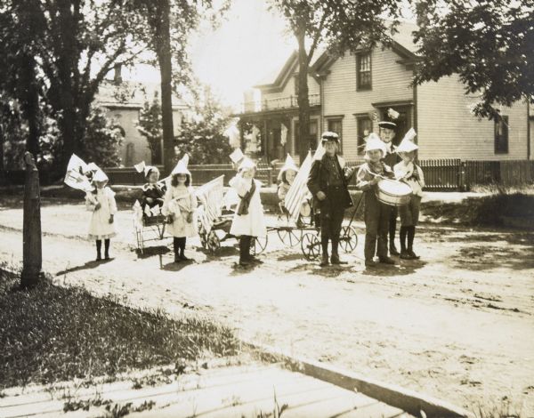 A group of children are parading down the street on the Fourth of July. The drummer is leading the way. Two girls and one boy are pulling carts with a child in each cart. All of the marchers in the parade are wearing festive hats and/or carrying flags. There is a large wood frame house in the background. Caption reads: "Fourth of July Parade."