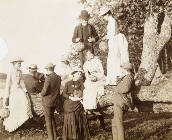 A group of young men and women are visiting McCauslin Brook. Standing on the tree trunk, W.A. Holt is holding a paper lantern. The woman, standing next to W.A. Holt, also has a paper lantern in her hands. The woman sitting on the log in front of W.A. Holt has a paper lantern in her lap. On the far left, a woman, possibly Ellen Holt, is holding a paper lantern too. The woman, wearing a dark outfit in the center front, is cradling a basket of fruit. A man, on the left, is pointing in the distance with his umbrella. Photo album page heading reads: "McCauslin Brook."