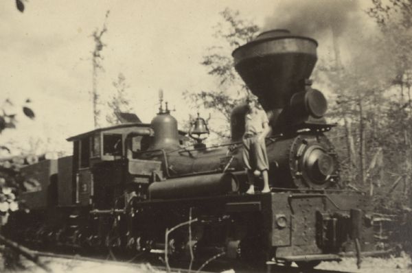 View towards a man standing on the left front side of a locomotive with his hands in his pockets. Smoke is spewing from the train's smokestack. There are large logs on the railroad car just behind the locomotive. Page caption reads: "At the Camp."