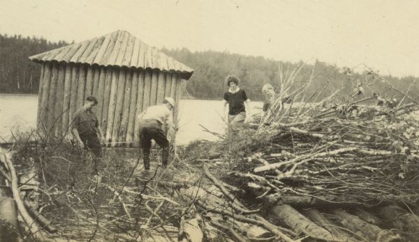 A small group is filling in part of the Archibald Lake shoreline with branches to protect the point from erosion. Names from left to right: Pete Blanchard, W.A. Holt, Juliet Stroh, and Lucy Holt. A small building made of split logs is behind the group. Caption reads: "Filling in to Protect the Point."