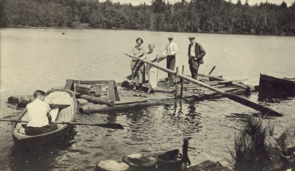 W.A. Holt and several visitors are paddleboarding or rafting on Archibald Lake. Names from left to right: Catherine Wilson, Nora Klass, W.A. Holt, and George Bangoff. Donald Holt is rowing a clinker-built duck boat toward the raft. The pier is in the foreground.

Caption reads: "Miss Catherine Wilson of Jacksonville, Florida, Miss Nora Klass, W.A.H., and Mr. Bangoff."