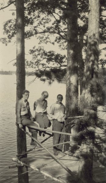 Three young adults are on the deck over looking Archibald Lake. Tall white pine trees are visible. Names from left to right: {Unknown}, Lillian Wheeler (?), and Donald Holt.