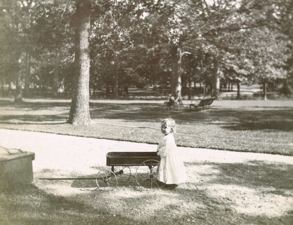 Alfred Holt is leaning on a toy wagon with wooden wheels at grandmother Holt's home, which was known as "The Homestead." The address of the Devillo and Ellen Holt home, a symmetrical Italianate villa, was 570 N. Sheridan Road in Lake Forest, Illinois. The page heading reads: "At Grandmother Holt's - Alfred," and the photo caption reads: "Wagging."