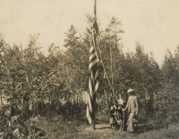 The American flag, twisted around the wooden pole on the Island, is the focal point this picture. Lucy and W.A. Holt with their children are standing to the right of the flag pole. Jeannette Holt seems to be doing the honors of raising the American flag. Alfred Holt, with his hands on the rope, is standing to the left of Jeannette Holt. Toddler Eleanor Holt is standing in front of her father, who is also holding a rope. The letters "SL," part of the Island Lodge flag, can be seen above the American flag on the pole. There are tall trees in the background.