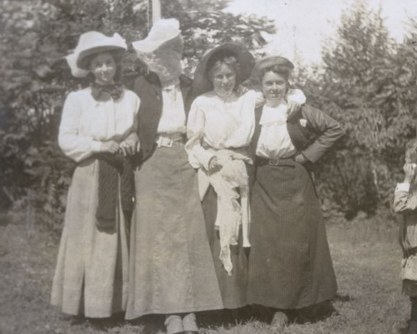 A portrait of four young women standing close together before going hiking. Judging by their body language, these young women appear to be pals. They all are wearing hats and skirts. One woman (second from left) is wearing netting over her face and leaning against the flag pole behind her. There are trees in the background. The caption reads: "Ready for a Tramp."

 