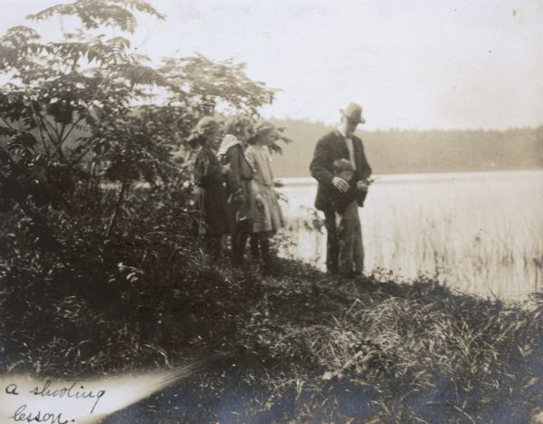 Grandfather I.P. Rumsey is teaching grandson Alfred Holt how to shoot a gun on the shore of Archibald Lake. Jeannette Holt and the Stroh girls are watching the shooting lesson. The caption reads: "The Shooting Lesson."