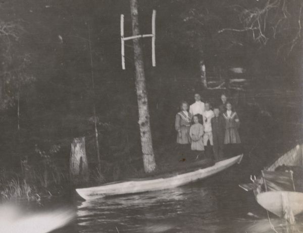 View across water towards members of the Reading Circle, including one woman and six children. The "H," made of birch branches, marks the location as being Hermit Island. Two boats are sitting on the shore of Archibald Lake. Caption reads: "Reading Circle at the Hermit." 