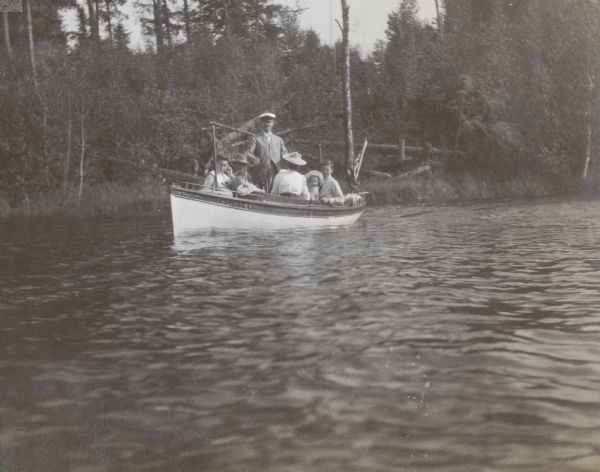 Standing in the center of the <i>Islander</i> steam launch on Archibald Lake, W.A. Holt is wearing a sea captain's hat. Friends and family are going with W.A. Holt to the Landing where they will see him off after two weeks of vacation on the Island. Names from left to right: Harriet McClure, unknown man, Captain W.A. Holt, unknown woman with back to the camera, Eleanor Holt, and James McClure. Trees and shoreline vegetation are in the background. Caption reads: "Departure of W.A. Holt after Two Weeks."

