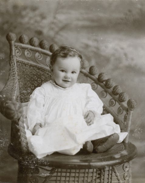 Studio portrait in front of a painted backdrop of Alfred Holt sitting in a Victorian wicker chair. He is looking at the camera and smiling. Caption reads: "July 1898 - Alfred."