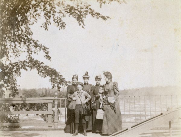 Standing with the Menominee River in the background, Mary Mathilda Axtell Rumsey and her family posing for a picture. Names from left to right: Mary Mathilda Axtell Rumsey, Wallace Rumsey, Minnie May Rumsey, Juliet Rumsey, Juliet Lay Axtell or Harriet Tracy Axtell. Wallace Rumsey is carrying a camera case. Caption reads: "Seeing Niagara - Mother, Minnie, Juliet, Auntie, Wallace."