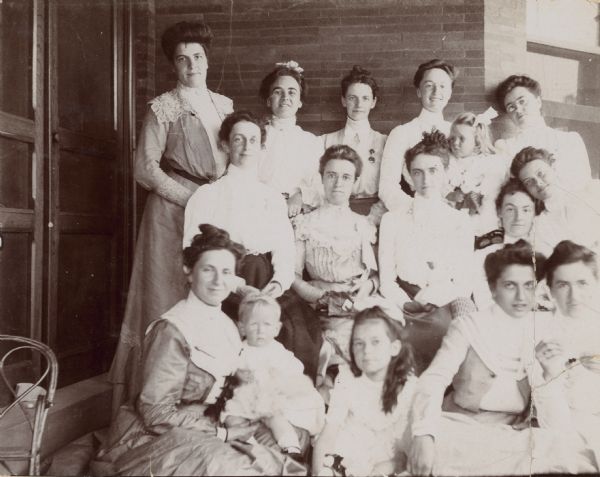 A portrait of a group of women outside on the porch, with a brick wall in the background. They are attending a reunion luncheon in Milwaukee. Lucy Rumsey Holt is standing in the center of the back row. There are several children in the picture. Caption reads: "Reunion Luncheon in Milwaukee." 

