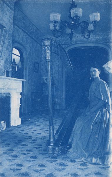 Lucy Rumsey is sitting with her concert harp in a home setting. She appears ready to play. There are arched doorways, a chandelier, and a fireplace in the background.