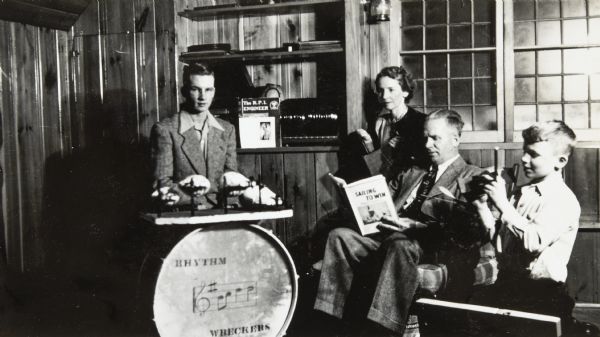 Doug DeWitt is sitting behind the drums. The words "Rhythm Wreckers" are printed on the drum face. Behind Doug DeWitt, on a shelf, are several things: a record player, records, a copy of "The R.P.I. Engineer," and a military portrait. To the right are Doug DeWitt's parents Eleanor Holt DeWitt and Donald S. DeWitt, who is holding "Sailing to Win" by Robert N. Bavier. Robert (Bobby) DeWitt is playing with a wooden toy, on the far right. Wood paneling and a window are in the background.