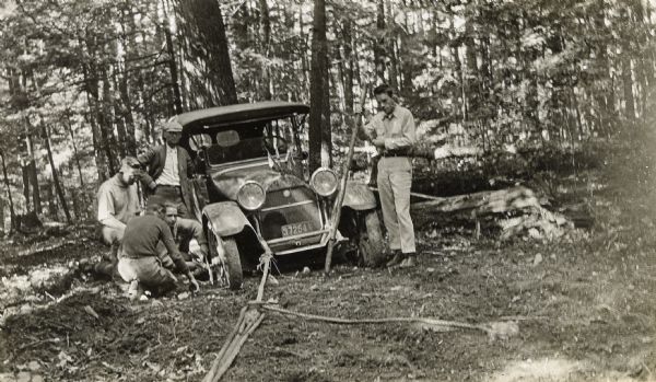 W.A. Holt is standing next to his broken down car in the woods. Three men on the left are examining the wheel of the car. One man is holding a crowbar. On the right, a man is standing and holding onto a long stick near the front wheel. In the foreground is a rope and other equipment tied to the front bumper. Tall trees are in the background.
