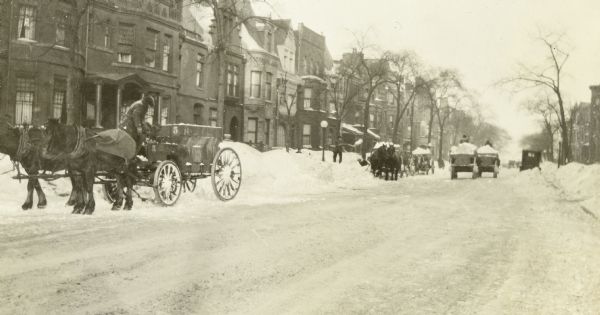 View down street towards men clearing snow from a residential street using shovels and horse-drawn wagons. A row of homes is on the left. Further down the block a car is parked along the right curb.