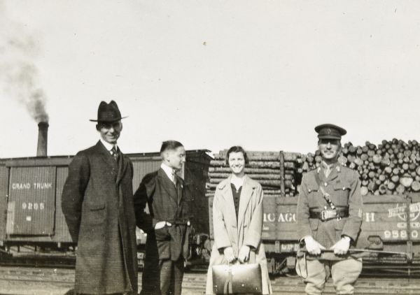 Captain Edwards (r) is standing at the train station with from left to right: W.A. Holt, Alfred or Donald Holt, and a woman, possibly Madeline Wood. In the background are two railroad cars. The car on the right is carrying logs and has the words "Chicago" and "Northwestern Line" painted on its side. Behind the railroad cars is a smokestack.

