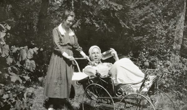 A man is dressed up as a big baby and is sitting in a wicker baby carriage. He is drinking from a glass bottle. A woman wearing round eyeglasses is standing behind the baby carriage.