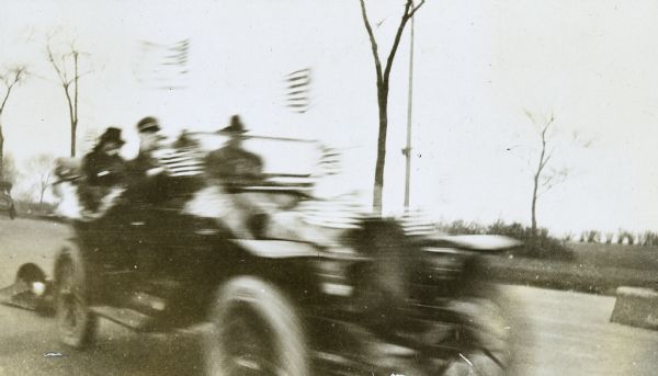 A driver and three or four passengers are in a moving car with the top down. Many American flags attached to the car are flying in the wind. The group may be celebrating the end of WWI.