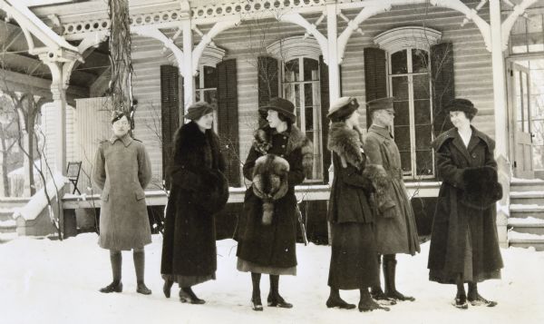 Wearing his cap, overcoat and boots, Donald Holt is looking at the camera. He is standing by himself on the left with his hands behind his back. Next to Donald Holt, Margaret Stroh and Juliet Stroh are talking while warming their hands with fur hand mufflers. On the right, Harriet Stroh, Alfred Holt, in uniform, and Elizabeth Stroh are standing together. These two women are also using fur hand mufflers. Two of the four women are wearing mink stoles. Snow is on the ground. In the background is a wooden building with a porch and tall arched windows with shutters.