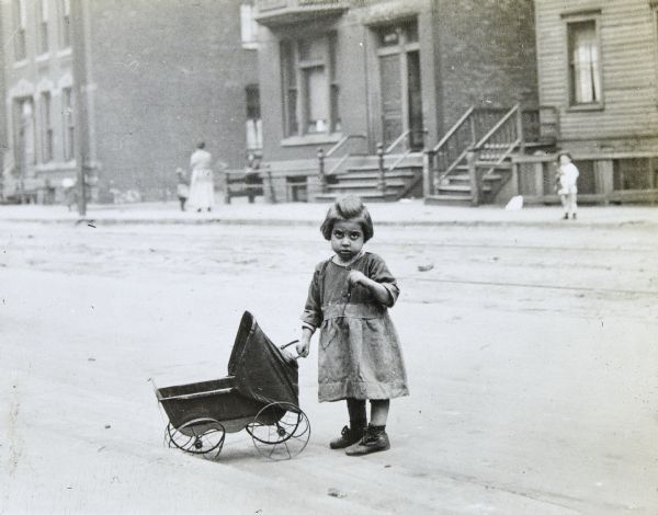 A girl is standing in the street near the curb with a baby carriage. She is wearing a dress and shoes. A woman and two children are standing on the sidewalk across the street.