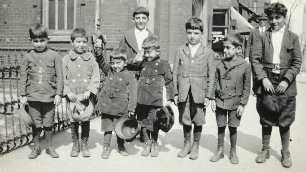 Young school boys are lined up in a row on the sidewalk. An older boy is standing behind the group. The young boys are all wearing short pants. One boy (third from the left) is raising his arm and holding a pack of cards. Another boy (fourth from the left) is saluting. An iron fence and a brick building are in the background.