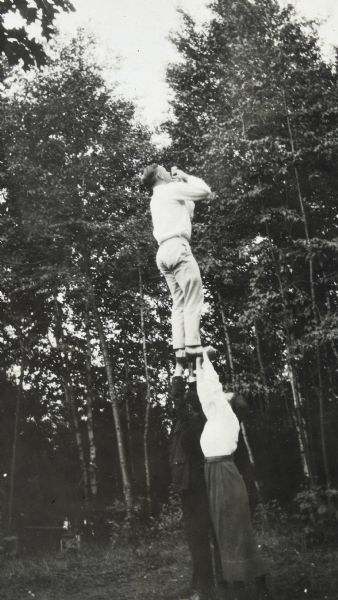 A man and woman are holding up a man who is balancing above them on their hands. He is holding his hands to his mouth. Tall trees are in the background. Page caption reads: "Summer 1919."