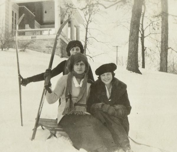Three young women are posing for a winter portrait with a sled and wooden skis. There is a building with a porch on a hill in the background.
