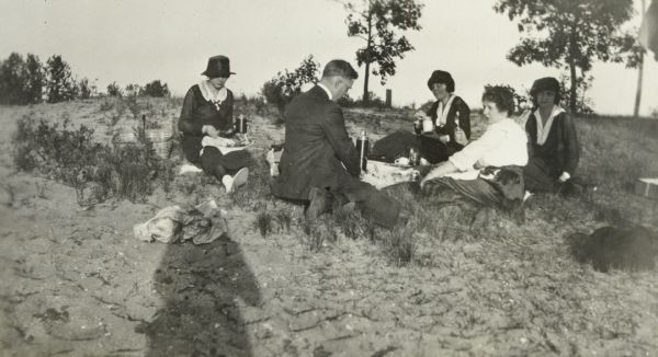 A group of one man and three women is picnicking on a grassy patch of sandy beach. The group is sitting on the ground and eating around a blanket with a Thermos on it. Page caption reads: "The Howards at Kalamazoo." The photographers shadow is in the foreground.