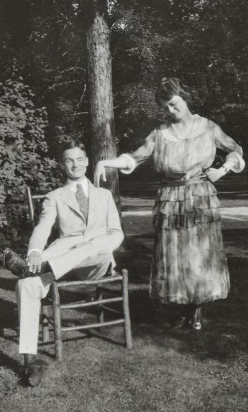 Alfred Holt and Madeleine Wood are outside on a sunny day. She is standing and holding a pose that suggests dancing. He is sitting in a wooden chair with his one foot crossed over his knee. Alfred Holt's eyes are either closed, or he is squinting in the sunlight. Madeleine Wood is looking down.

This outdoor portrait seems to be a spoof on the traditional portrait of a newly married couple. There is a matching photograph in the album of Alfred Holt and Madeleine Wood looking prim and proper.

Page caption reads: "July 1920," the month in which Alfred Holt and Madeleine Wood were married.