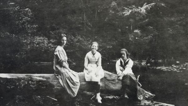 From left to right: Eleanor Gibson, {unknown}, and Helen Macartney are sitting together on a large fallen tree trunk partially submerged in Archibald Lake. In the background are tall trees. Page caption reads: "At the Lake 1920."