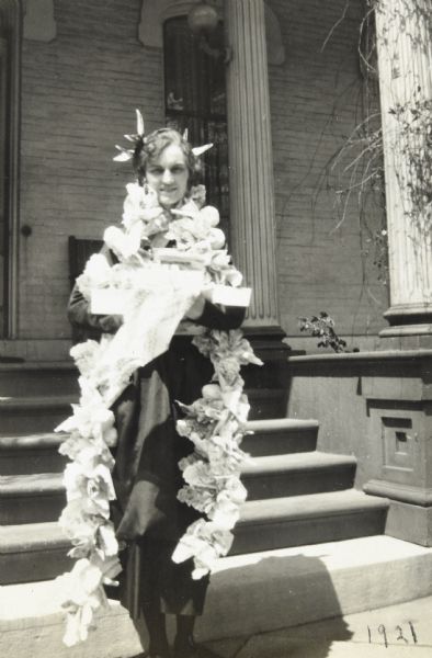 Celebrating her 25th birthday, Jeannette Holt is dressed for the festivities, wearing decorations in her hair, and paper flowers draped around her neck. She is standing and holding a gift box at the base of a set of stairs at The Evergreens, the Rumsey estate. Caption reads: "Birthday in Lake Forest, May 8, 1921." 