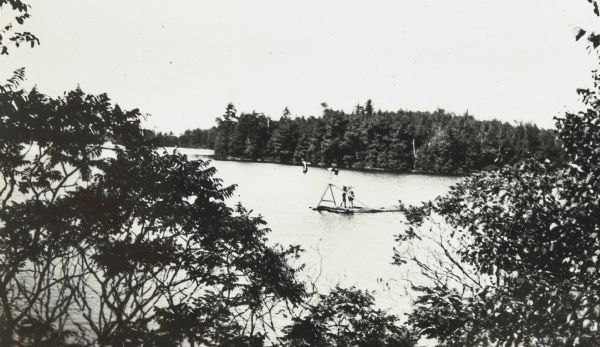 Elevated view through trees towards a man preparing to dive or jump off a wooden diving board, mounted on a barge, into Archibald Lake. Waiting his turn, another man is bent over at the top of the sloping ladder behind the diving board. There are three other people on the barge. Page header reads: "Summer 1921."


