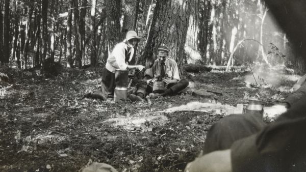 With her face partially shaded by a hat, Jeannette Holt is holding a cast iron frying pan sideways to shade the camera as a young man, sitting at the base of a tree, prepares to shoot. Her elbow is resting on a stack of metal containers. The camera, held by the young man, is resting on a bucket and is aimed towards the photographer. In the foreground on the right is the shoulder of a person sitting on the ground near a picnic blanket with food and tableware on it. A campfire is smoldering just beyond the blanket. A forest is in the background.