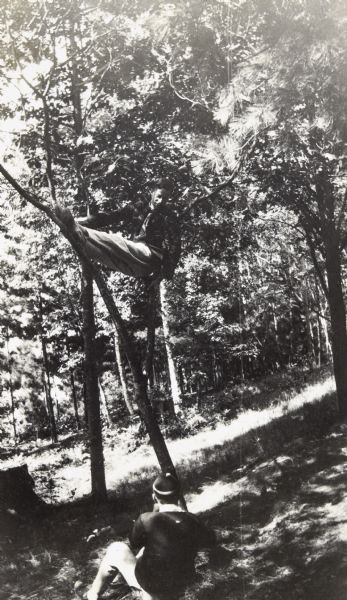 With legs extended, Lowell Hoxie is sitting up high in a tree, supported on two branches as if he were in a hammock. Below is a young man, sitting on the ground, who appears to be taking a photograph of Lowell. Woods are in the background. Caption reads: "Lowell Hoxie." 