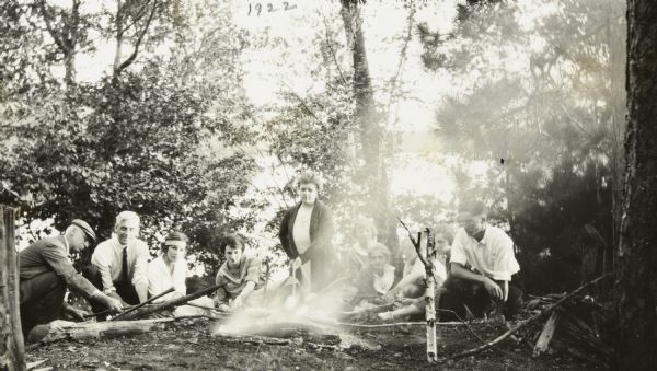 A group of Holts and Garrisons are roasting corn around a smoking campfire on the Island, with Archibald Lake in the background. W.A. Holt is on the far left and Mr. Garrison is second from the left. Jeannette Holt and Donald Holt are on the far right side. Garrison children and others are in the center. Page caption reads: "The Garrisons."