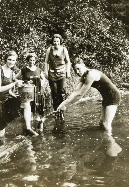 Jeannette Holt (far left) is standing in the water near the shoreline with one foot up on a log. She is balancing a metal pail on her knee, which has a screen and is filled with some kind of marine life, which the group presumably has collected. To the right of Jeannette are two Garrison women standing in the water. A Garrison man, standing on the right in the water, is leaning forward on the branch of a log floating in the water. Page caption reads: "The Garrisons."