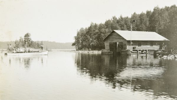 View of Henry Rumsey's boat house, which is built of logs. An American flag is flying from the flagpole at the front of the boathouse. 