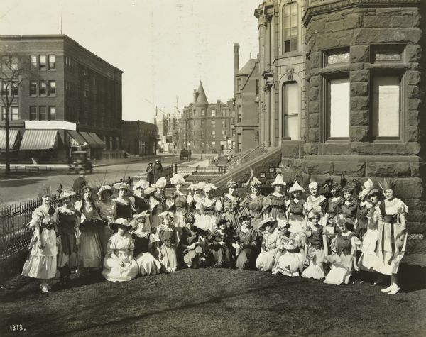 Outdoor group portrait of the Class of 1918 Flower Market at Gertrude House. In costume, Jeannette Holt is standing third from the left. She is holding a tray with flowering plants. This group of women is studying to be kindergarten teachers and immigrant settlement house teachers. In the background is Rush Street in Chicago.
