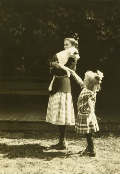 Jeannette Holt is standing and holding Spotty, the family dog, in her arms while facing to the left. Lillian Wheeler is standing behind her and reaching up to hold the paw of the dog, unseen by Holt. In the background is a wooden bench on a platform, surrounded by foliage.


