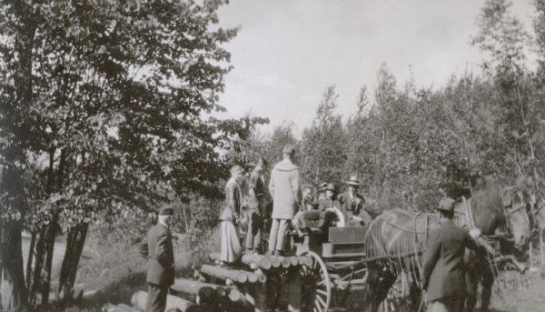 Juliet Rumsey Stroh is standing on a step leading up to the horse-drawn wagon, and three boys are standing on the top log step. A man and a woman are sitting in the front cushioned seat of the wagon. The empty seat next to them is reserved for the driver who is standing by the horses with one hand on the harness. There are two or three people in the back seat of the wagon. On the far left, I.P. Rumsey, wearing a cap, is watching the impending departure of the guests. Trees are in the background.